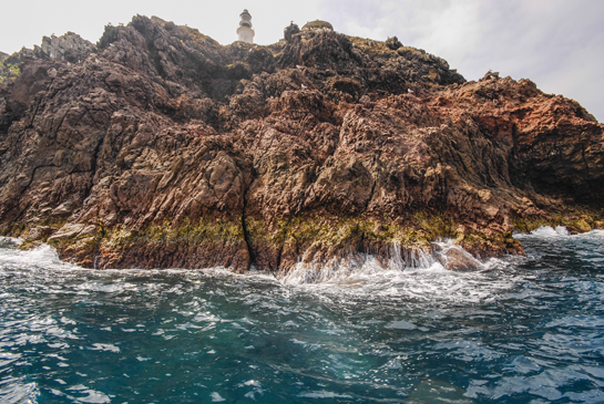 Vista general del punto de inmersión, en la parte superior se puede ver el faro. Se trata de una de las zonas mejor conservadas, desde el punto de vista de las comunidades submarinas, debido a la dificultad de acceso por tierra, que reduce la pesca desde la costa, y a la vigilancia que realiza el destacamento militar desde la Torre de la Conquista, que impide la pesca artesanal por parte de los pescadores marroquíes.